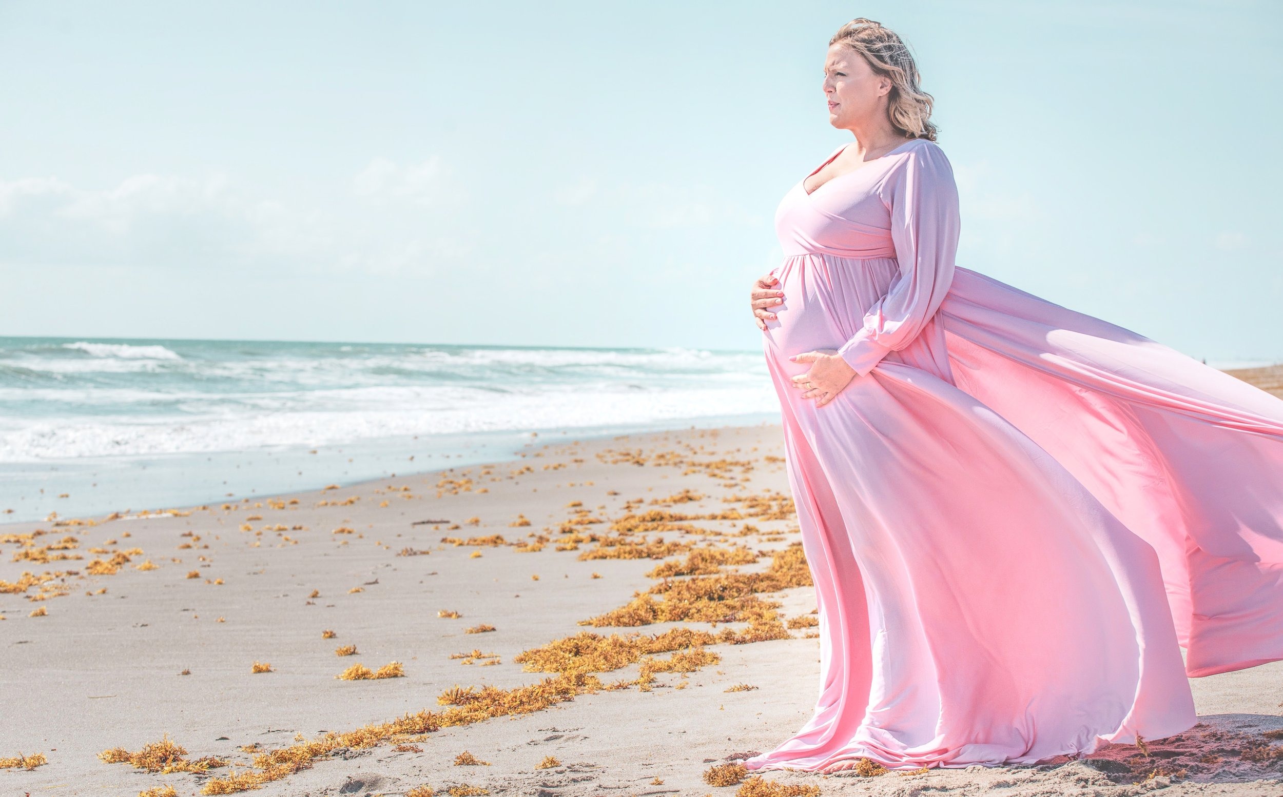 An expectant mother walking beneath a soft pastel sky, waves humming at the shore below her.