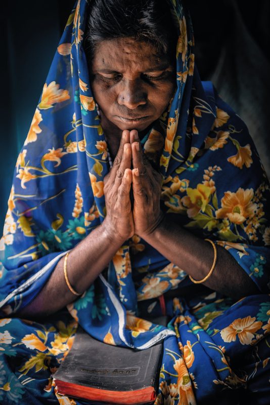 A woman with hands clasped and a Bible near, praying quietly as the world around her fades away.