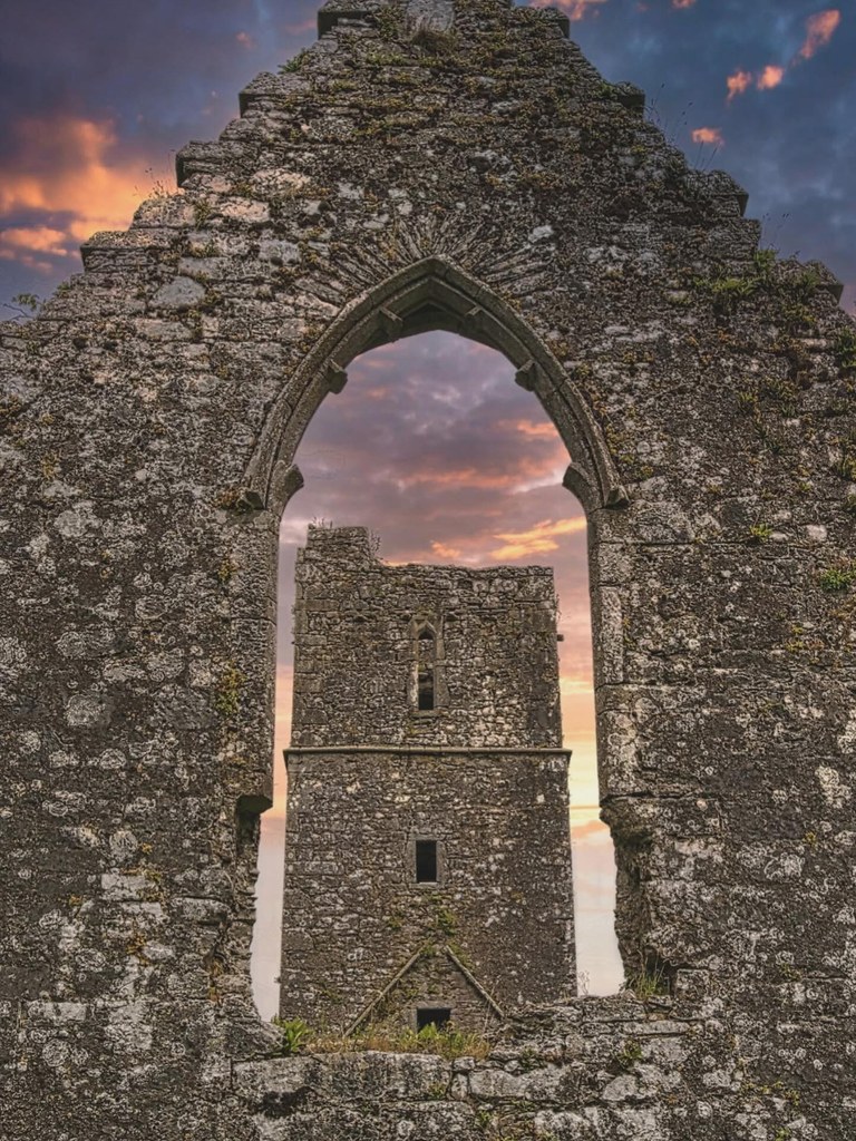 The ruined Kilcrea Abbey framed through a stone arch at sunset, the sky burning orange above the old friary walls.