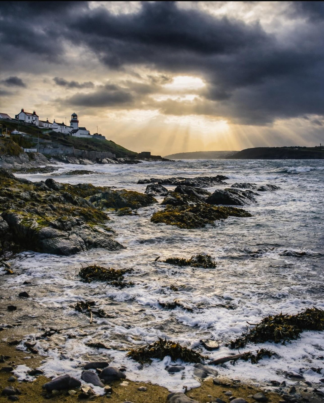 A lighthouse stands against a breaking storm, shafts of light cutting through heavy cloud over a boiling sea off the Cork coast.