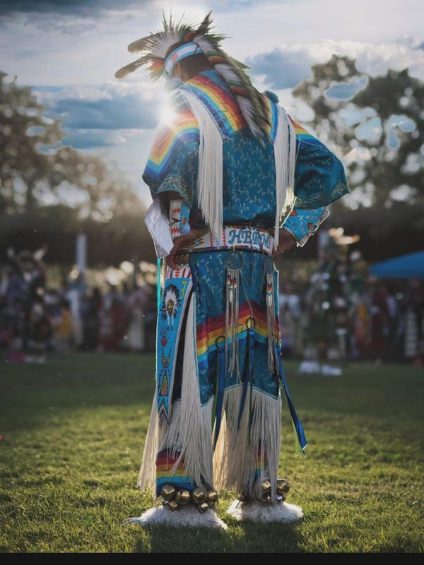 A powwow dancer seen from behind, backlit, feathers and regalia rimmed with light.