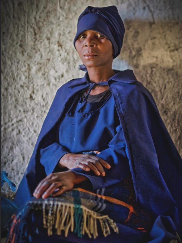 A woman in a vivid blue sari carrying a basket through a narrow lane, early light cutting through dust and smoke.