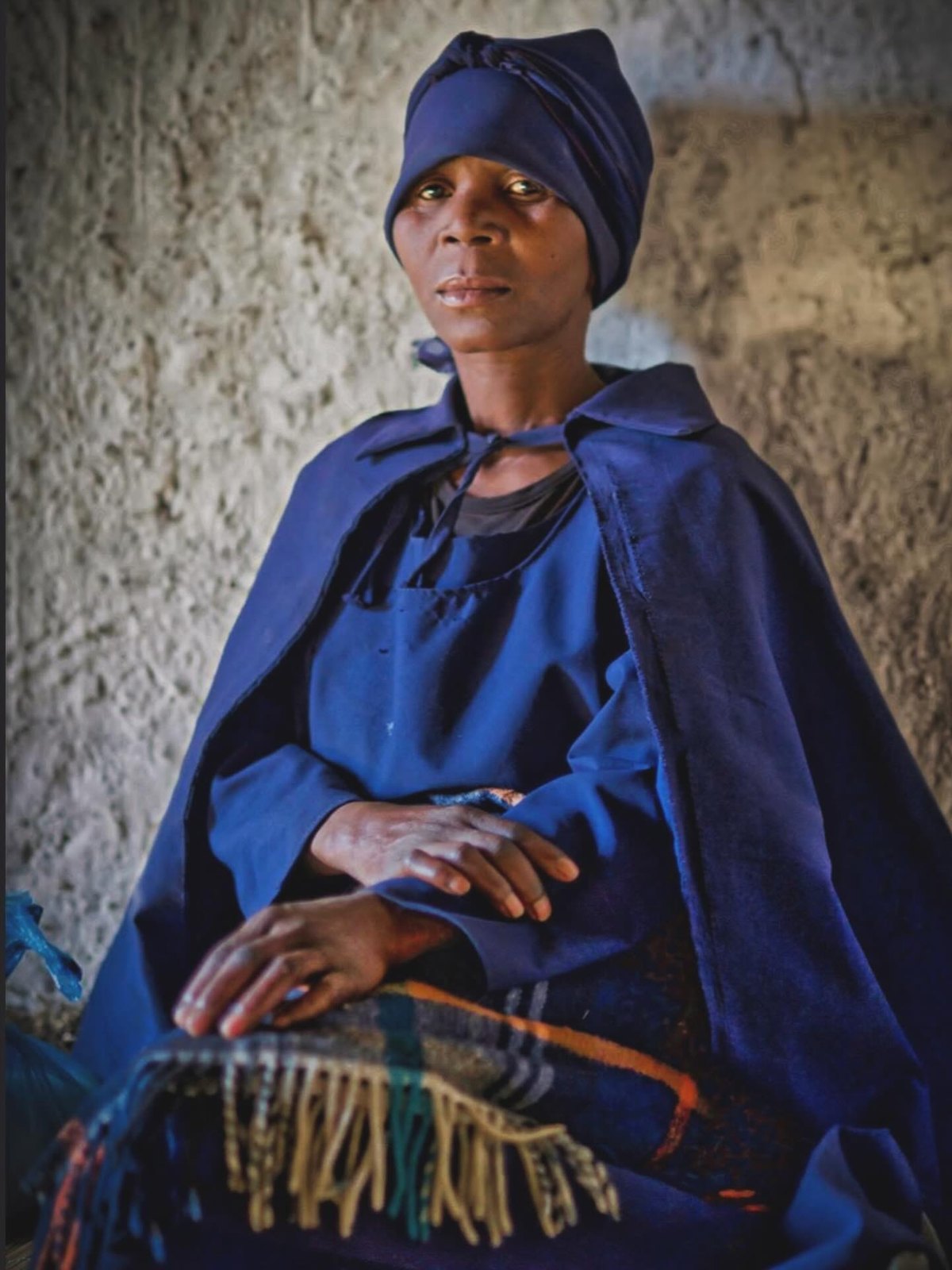 A woman in a vivid blue sari carrying a basket through a narrow lane, early light cutting through dust and smoke.