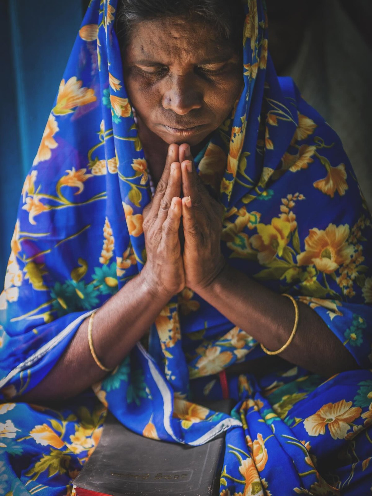 An Indian woman in a blue floral sari with hands folded and eyes closed in prayer, faith held quietly and inwardly.